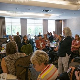 Une foule diversifiée de personnes assises et écoutant une femme debout avec un microphone au milieu de la salle lors du forum Inclusive Housing Solutions Lab au Nouveau-Brunswick.