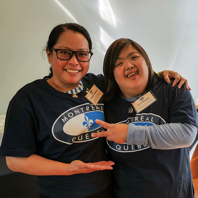 Deux femmes de L'Arche Calgary portant des t-shirts bleus sourient, l'une d'elles faisant le signe de la paix.